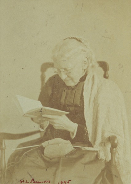 Elderly woman seated and reading a book with knitting in her lap, 1895.