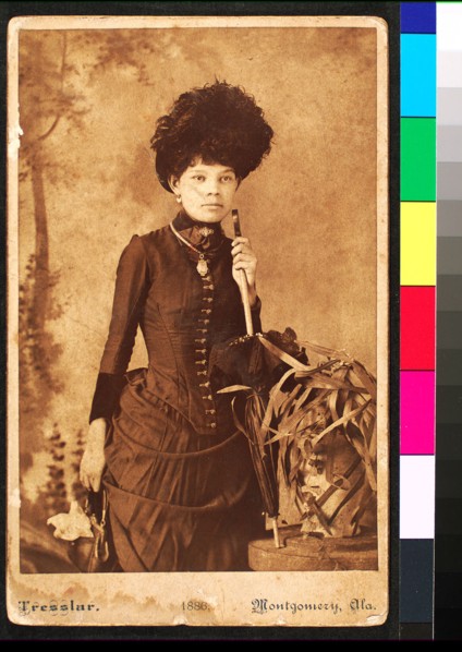 Standing woman, wearing dress with bustle, holding purse at side and umbrella resting..., 1886. Creator: Unknown.