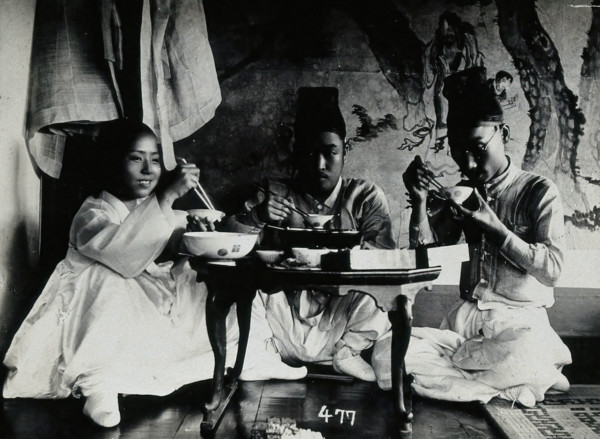 A group of people eating a meal, seated behind a small table, in front of a painted screen, c1900. Creator: Unknown.