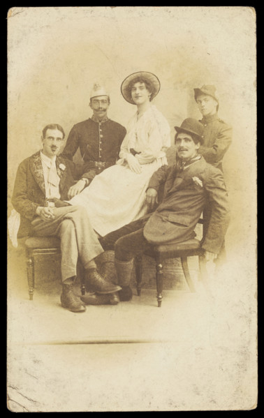 Amateur actors sitting down, surrounding a man in drag seated in the centre, [1910-1919?]. Creator: Unknown.