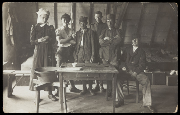 French prisoners of war, one in drag, gather round a small wooden table, [between 1910 and 1919?]. Creator: Unknown.