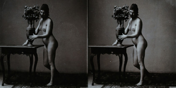 A young woman posing naked in a photographic studio, standing in front of a table on which..., c1900 Creator: Unknown.