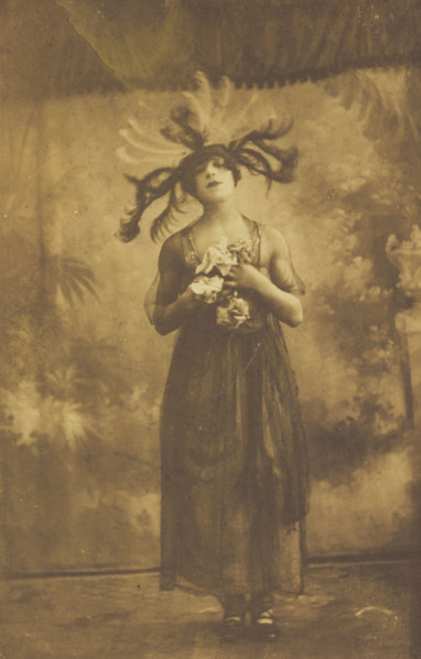 A performer in drag poses on stage wearing a head garment with sprawling feathers, [1910-1919?]. Creator: Unknown.