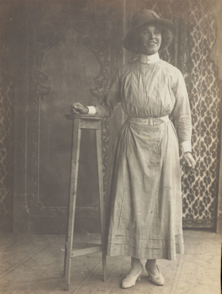 A sailor in drag leans on a high stool, in front of patterned scenery, (between 1910 and 1919?). Creator: Unknown.