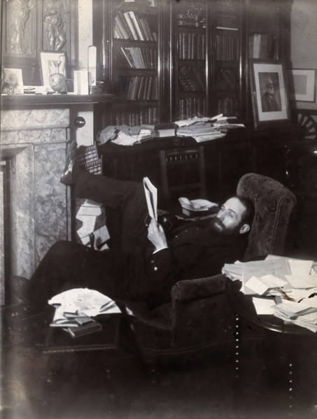 Arthur Schuster, lying on an armchair by a fireplace, with a photograph of Lord Kelvin..., c1901. Creator: Unknown.