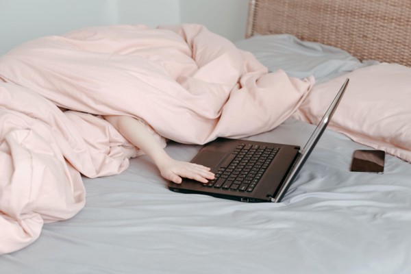Bored funny child lying in bed under blanket and learning in virtual online school class. Hands typing on computer keyboard. Kid working on laptop