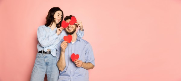 Flyer. Young and happy man and woman holding greeting cards shaped hearts isolated on pink trendy color background. Emotions, youth, love and