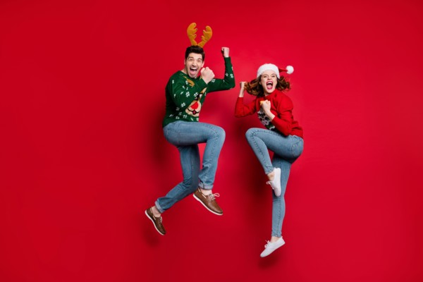 Full body photo of amazed jumping couple excited by x-mas prices wear ugly ornament jumpers and headwear isolated red color background