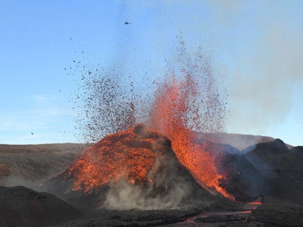 Volcanic vent at Fagradalsfjall, Iceland, erupting incandescent orange and red lava. Black lava in the foreground and blue sky in the background.