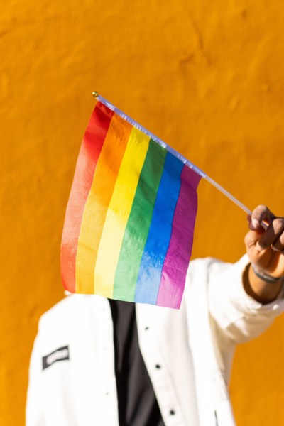 A young adult holds a multicolored rainbow flag in front of a bright yellow background
