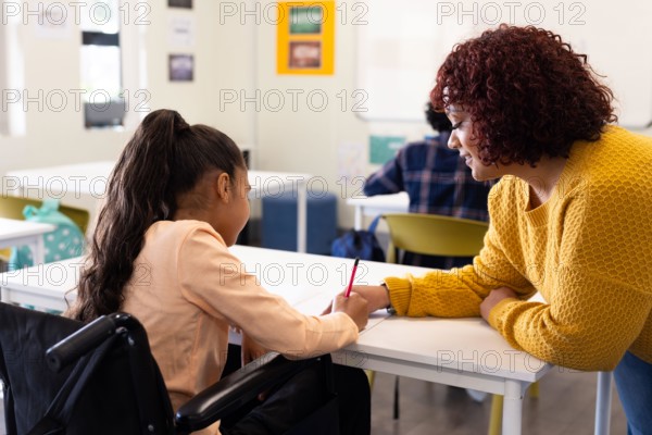 Female teacher assisting girl in wheelchair with writing task in classroom, at school