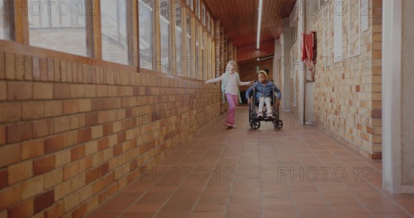 Two classmates rolling wheelchair and walking with backpack in school hall, with fire hose cabinet
