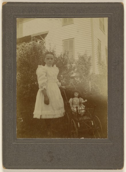 little girl in white dress standing next to a baby carriage with a doll inside; American; about 1900; Gelatin silver print