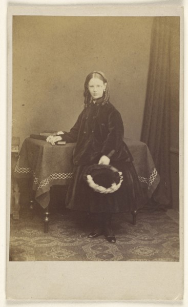 young girl in curls, standing, holding a hat, resting her other arm on a table with books on top; Joseph Brown, British, active