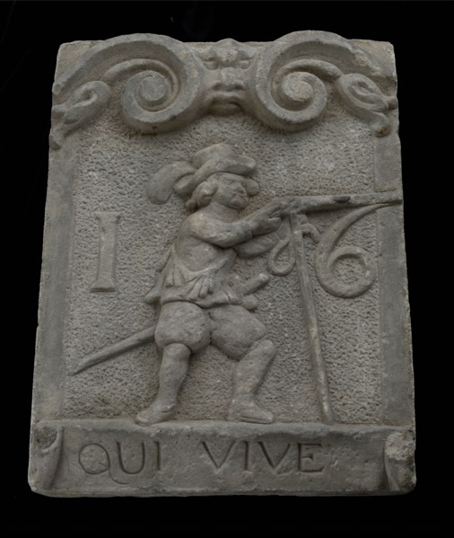 Wall brick from gate with musketeer and ribbon with text qui vive 16, facing stone sculpture sculpture building component