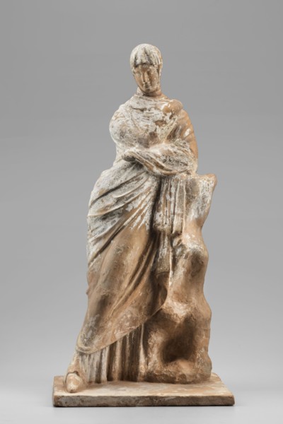 Female figurine. The figurine depicts a standing woman wrapped in a cloak, leaning her left elbow