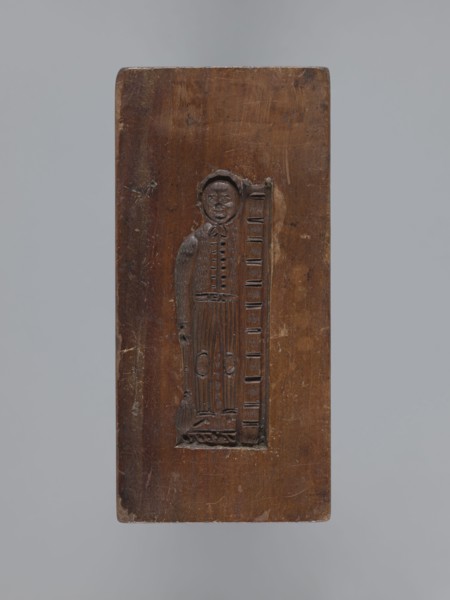 Double-sided gingerbread form. Side A: female figure with a scarf on her head, standing, leaning on