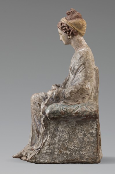 Figurine of a woman. A girl sitting in a chair is dressed in a soft white chiton over which a blue,