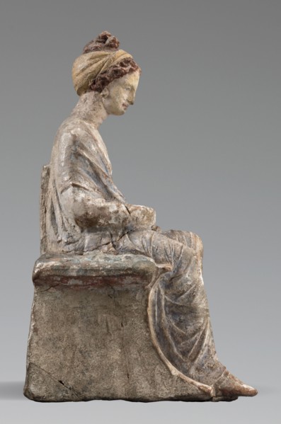 Figurine of a woman. A girl sitting in a chair is dressed in a soft white chiton over which a blue,