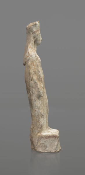 Female terracotta figurine of a standing woman, depicted frontally. Set on a small, hollow,