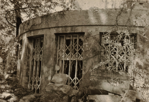 Circular part of a ruin, with bars in front of the windows, stones in the foreground and vegetation