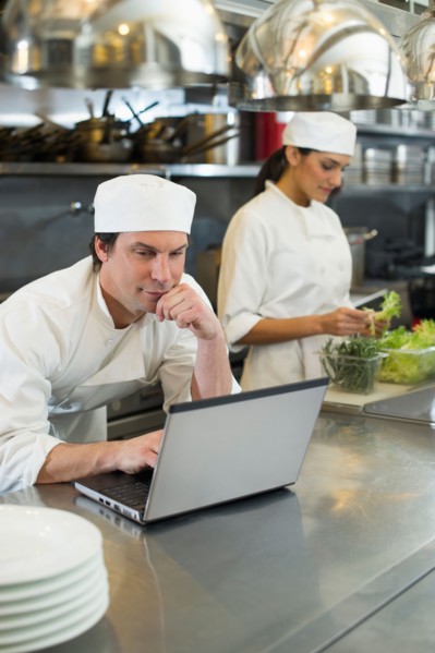 Chef using laptop and cook preparing food in commercial kitchen.