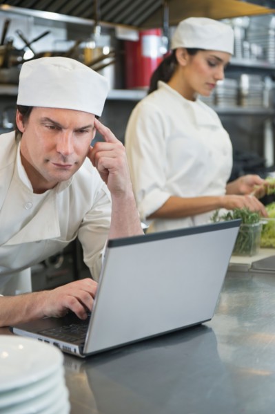Chef using laptop and cook preparing food in commercial kitchen.