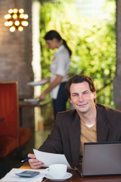 Owner working with laptop in restaurant with waitress in background.