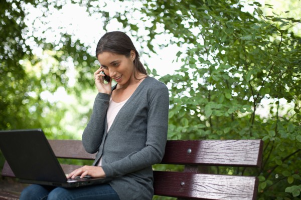 Young woman sitting on park bench using laptop and cell phone. Photo : Jan Scherders
