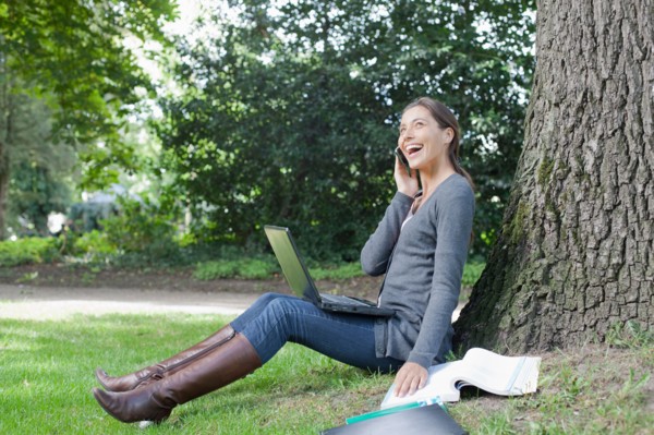 Young woman sitting under tree using laptop and cell phone. Photo : Jan Scherders