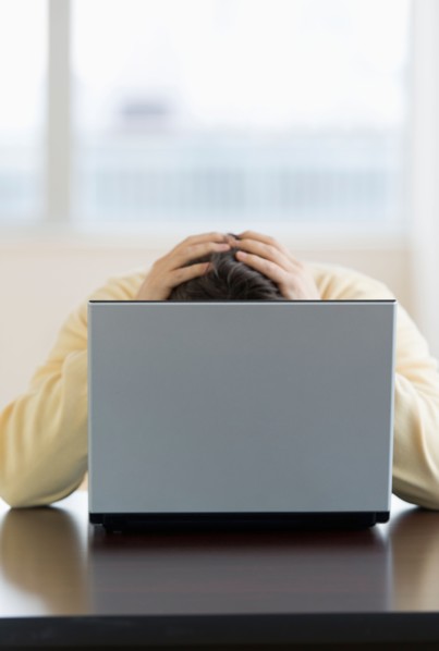 Stressed man sitting at desk behind laptop with head in hands.