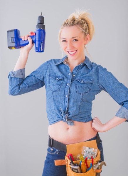 Portrait of young woman wearing tool belt and holding drill, studio shot. Photo : Daniel Grill