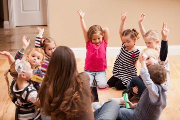 Children (2-3, 4-5) sitting on floor and raising hands. Photo : Mike Kemp