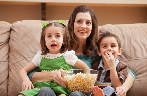 Front view of mother with children (4-5, 6-7) sitting on couch watching tv and eating popcorn.