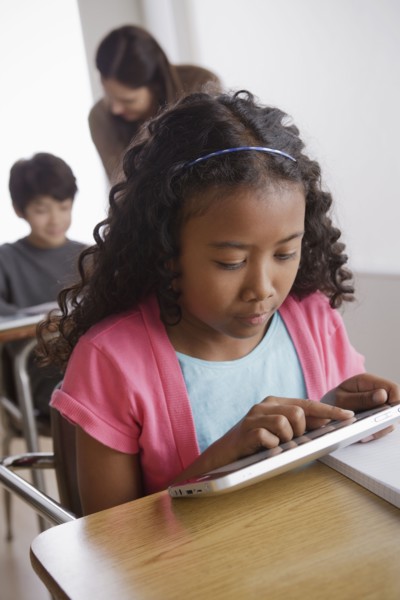 Schoolgirl (10-11) holding digital tablet with boy (12-13) and teacher in background. 
Photo : Rob