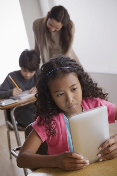 Schoolgirl (10-11) holding digital tablet with boy (12-13) and teacher in background. 
Photo : Rob