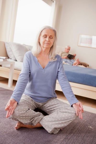 Senior woman practicing yoga with husband reading in background. Photo: Rob Lewine