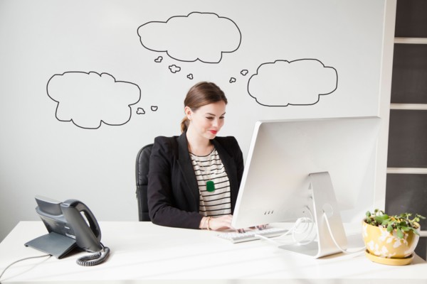 Studio shot of young woman working at desk with thought bubbles around her head. Photo : Jessica