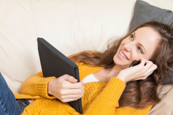 Woman lying on sofa with digital tablet and talking via cell phone