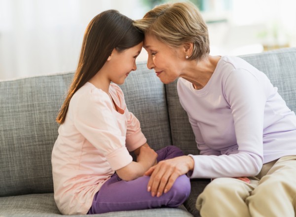 Granddaughter (8-9) and grandmother sitting on sofa and touching with heads.