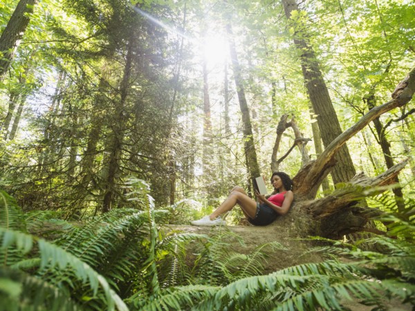 Young woman reading book on log in forest. USA, Oregon, Portland.