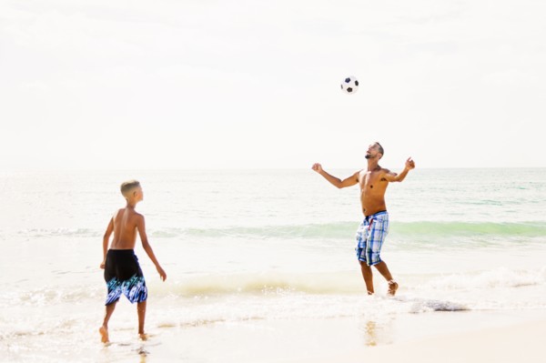 Father and son (10-11) playing soccer on beach