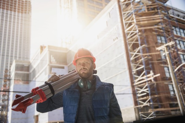 Construction worker carrying tripod on shoulder.