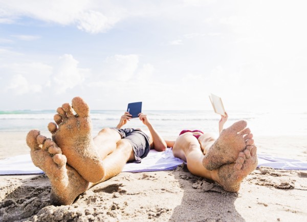 Close-up of sand-covered feet of young couple lying on blanket on beach reading