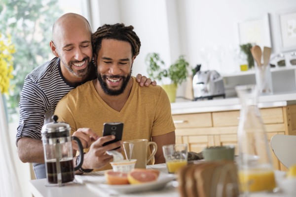 Smiley homosexual couple using smart phone at dinner table.