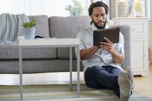 Young man with dreadlocks using digital table by sofa in living room.