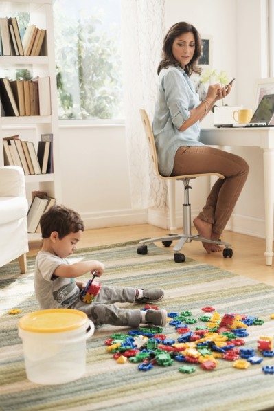 Boy (2-3) playing with toy blocks while mother working on laptop.
