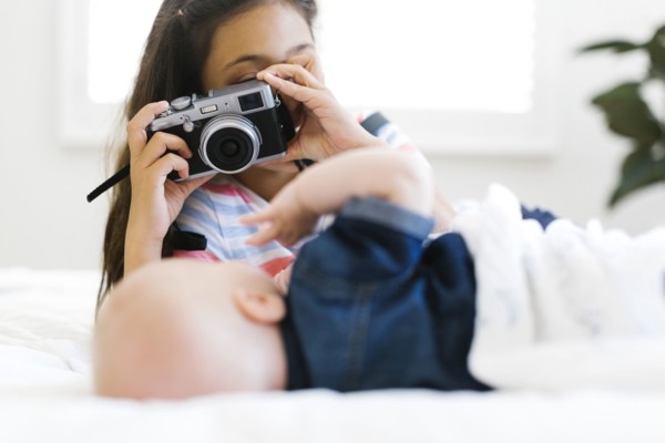 Girl (10-11) taking photo of her small brother (12-17 months) lying on bed