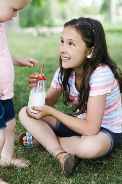 Girl (10-11) sharing bottle of milk with sister (2-3) in park