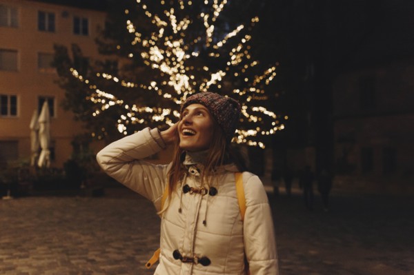 Smiling woman wearing coat by fairy lights in tree at night